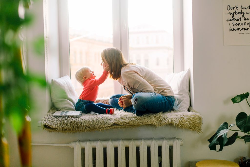 Baby reaching up to touch a woman's face. THey are seated in a window.