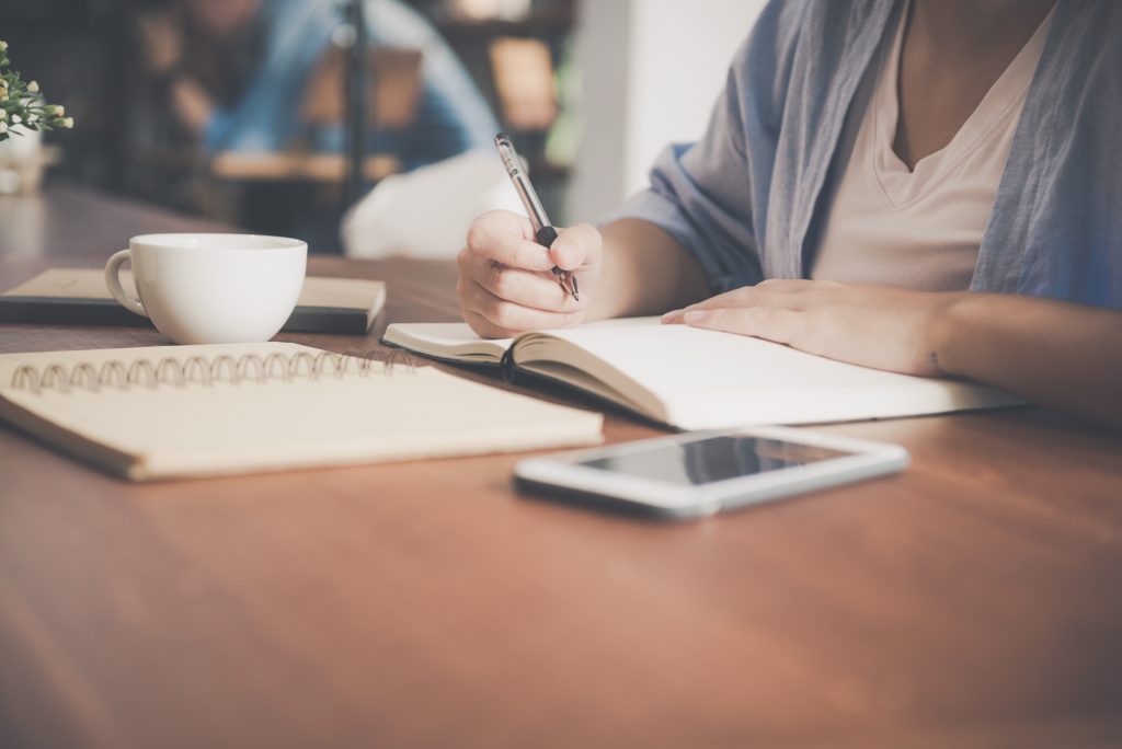 woman writing on a notebook besides teacup and tablet computer.