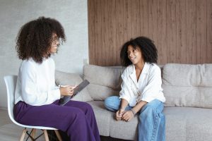 Two Black women talk to each other. One wears purple pants, glasses, and holds a clipboard which she’s writing on. The other wears jeans and sits casually on a beige couch.