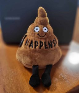 A brown poop shaped emoji plush sits on a table. On its front reads 'HAPPENS'.