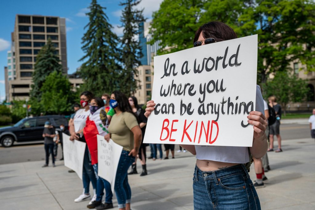 People standing and protesting with signs and face masks. One sign reads, "In a world where you can be anything, be kind."